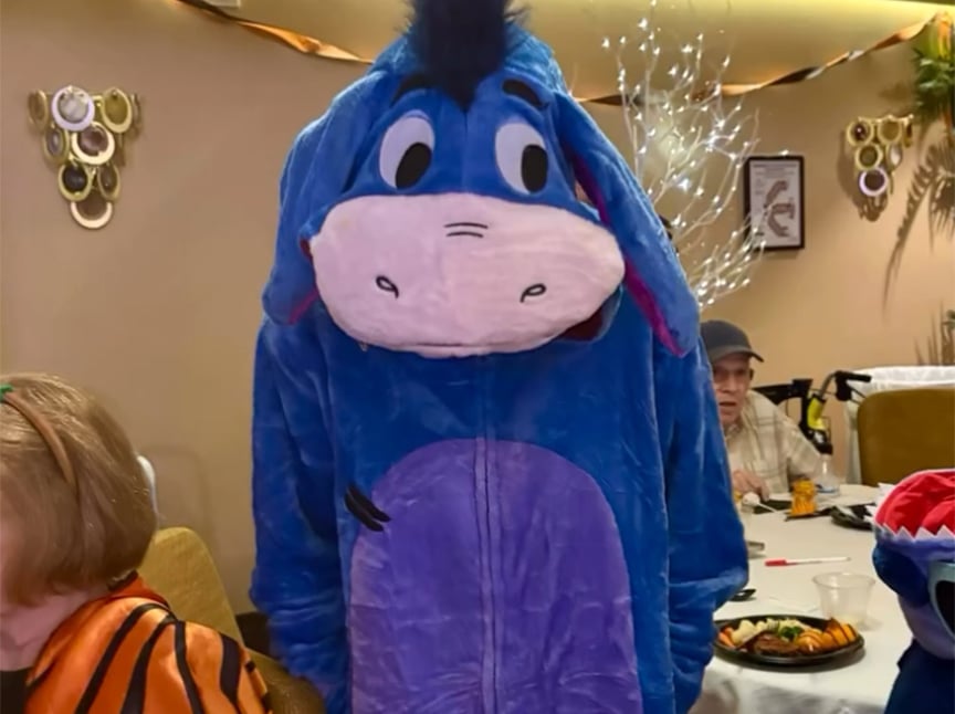 Screenshot Person wearing a blue Eeyore costume stands indoors at a decorated party with tables, food, and other people in the background. Orange and black streamers hang on the wall.
