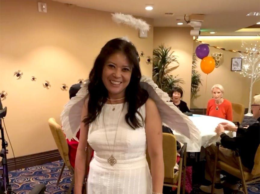 Screenshot A smiling woman in a white dress and angel wings stands in a decorated room with people seated at tables. Halloween decorations, including fake spiders on the wall and balloons, are visible in the background.