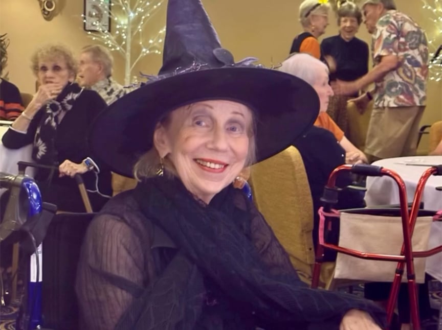 Screenshot An elderly woman smiles while wearing a large black witch hat and a sheer black scarf. She sits at a table with a walker nearby, surrounded by other seniors socializing at a festive indoor event.
