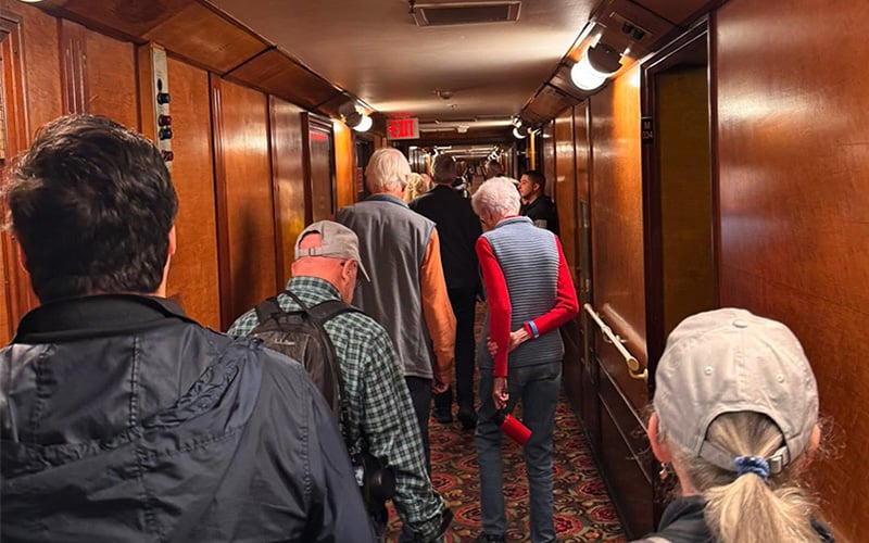 A group of elderly people walk down a narrow, wood-paneled hallway with carpeted floors, some wearing hats and jackets. The corridor is warmly lit with wall lights, and an exit sign is visible at the far end.