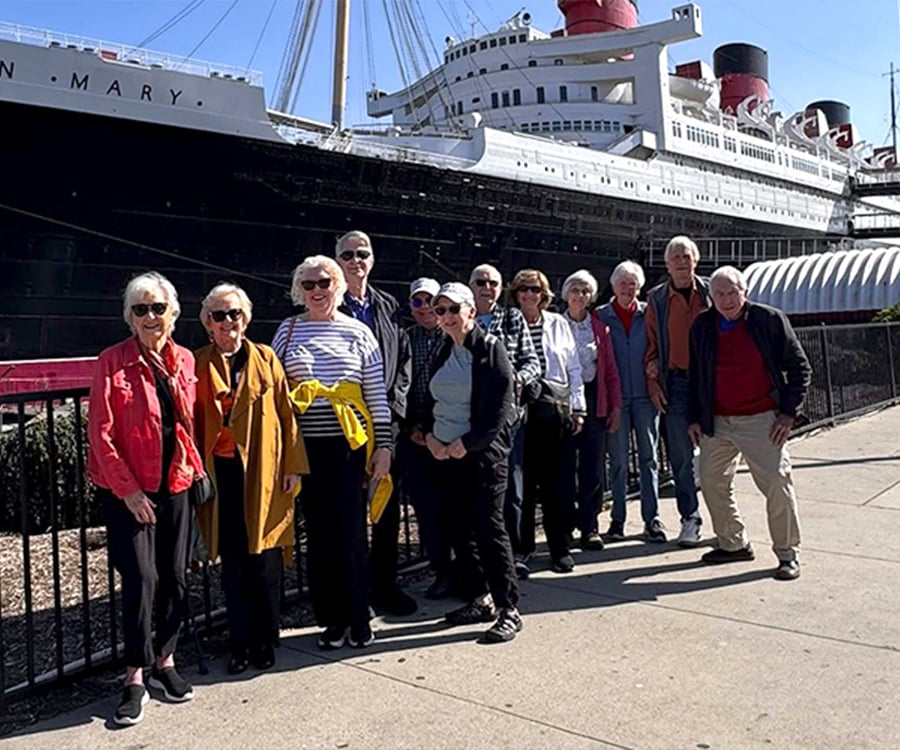 A group of twelve older adults pose and smile in front of the historic ocean liner Queen Mary, docked at a pier on a sunny day.