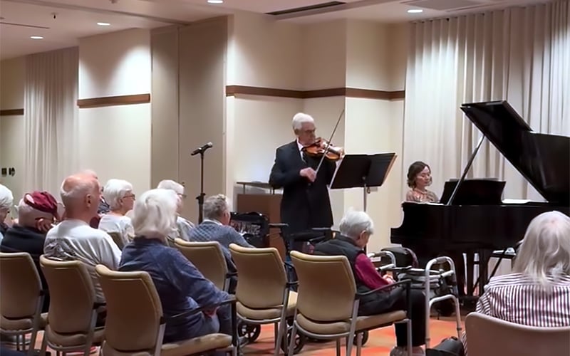 A man plays the violin and a woman plays the piano in front of an audience of elderly people seated in a room with beige walls and large windows covered by white curtains.