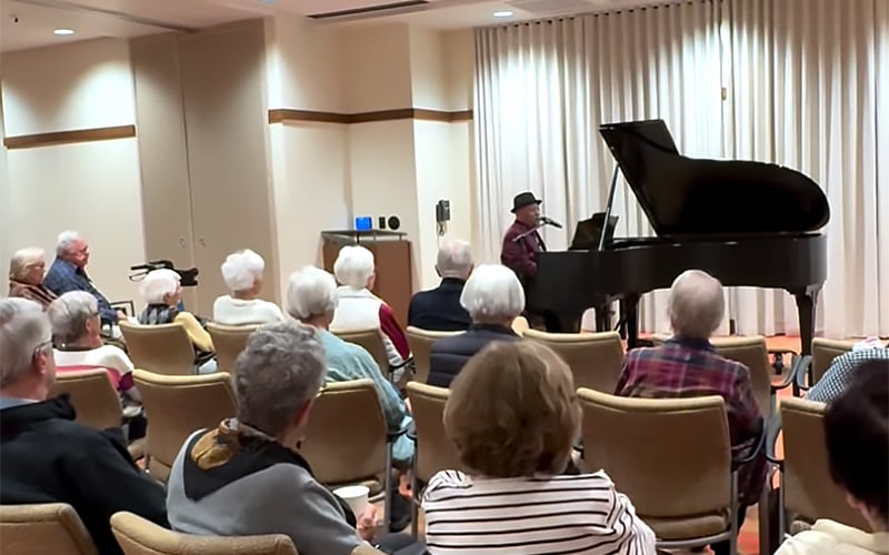 A group of elderly people sit in chairs watching a person in a hat play a grand piano in a room with beige walls and closed white curtains.