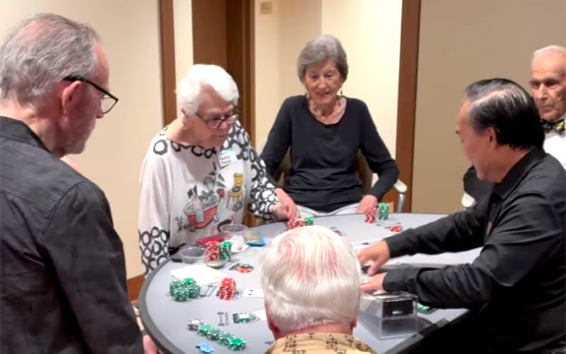 Five older adults sit around a circular table playing poker, with poker chips and cards spread out in front of them. They appear focused and engaged in the game.