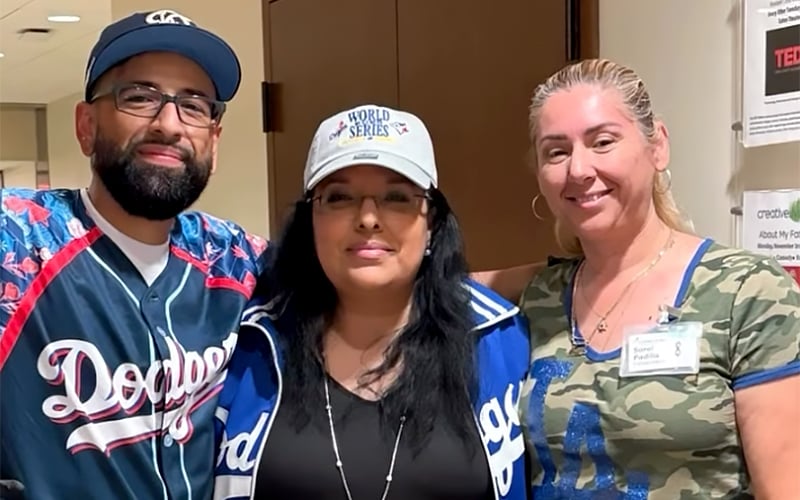 Three adults stand together indoors, smiling at the camera. The man on the left wears glasses, a cap, and a Dodgers jersey. The woman in the center wears a World Series cap and jacket. The woman on the right wears a camouflage shirt.