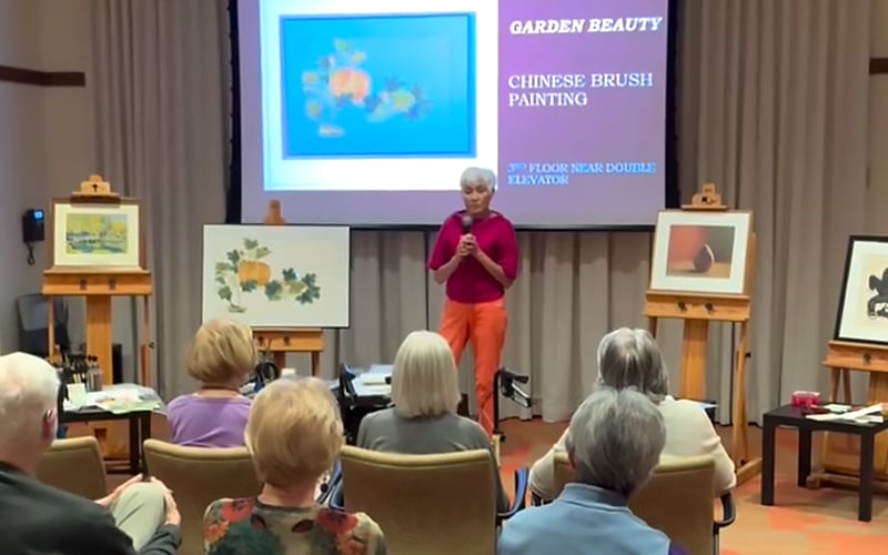 A woman stands in front of an audience, speaking at a presentation on Chinese brush painting. Several paintings are displayed on easels, and a slide behind her reads Garden Beauty Chinese Brush Painting.