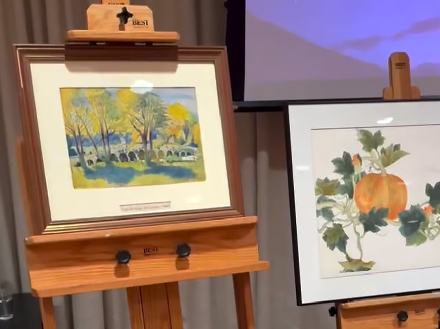 Two framed paintings on wooden easels: the left one depicts trees and a stone bridge in bright colors, and the right one shows a pumpkin with green leaves. A curtain and part of a screen are visible in the background.