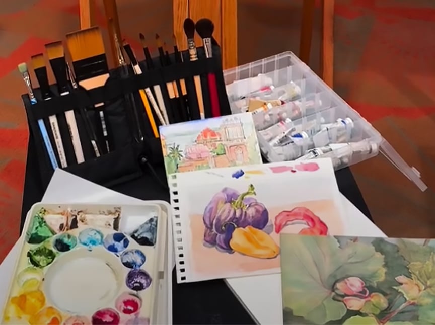 A table with watercolor paints, brushes, a palette, a plastic case with paint tubes, and several watercolor paintings featuring buildings, fruit, and a flower.