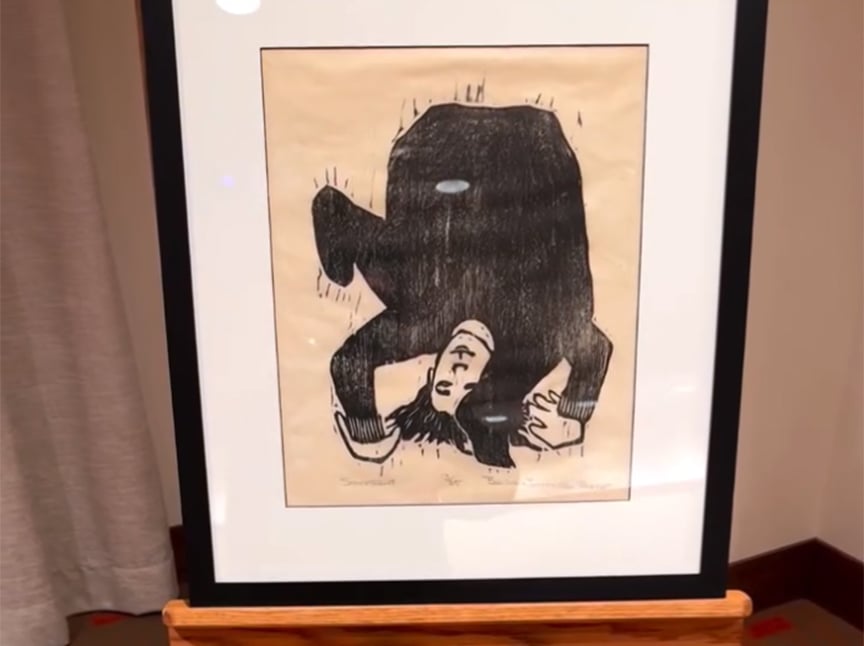 A framed artwork on an easel shows a person in dark clothing curled up upside down, with their face and hands visible, against a plain background. The picture is displayed indoors near a beige curtain.