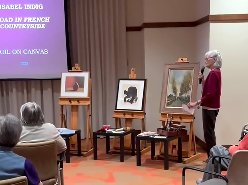 A woman stands with a microphone and paper, presenting to an audience in a room. Three paintings on easels are displayed beside her and a projection screen is visible with text about an artist and their work.