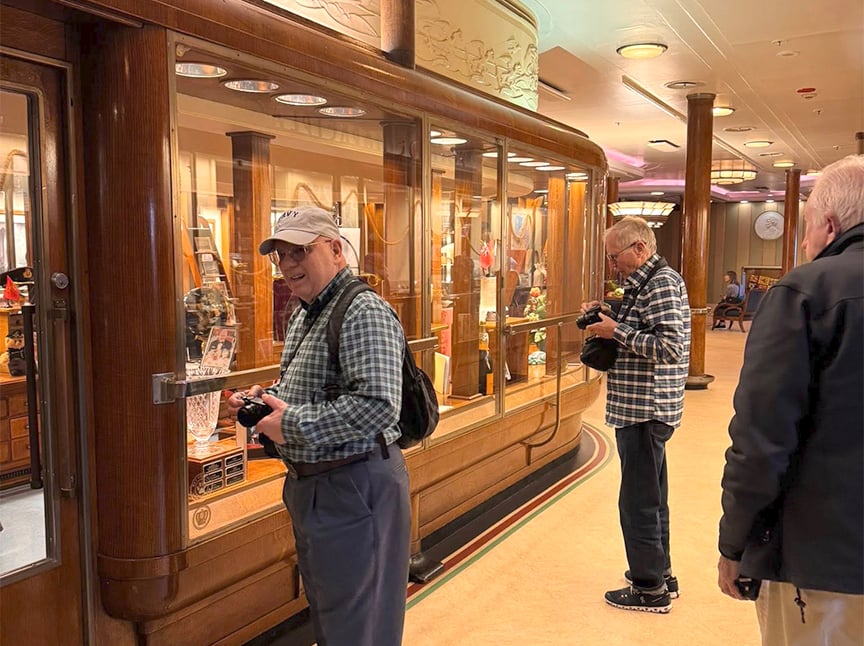 Three older men stand in front of a large, polished wooden display case filled with various objects. Two hold cameras and appear to be taking photos, while the third looks at the display. The setting appears to be a museum or exhibit.