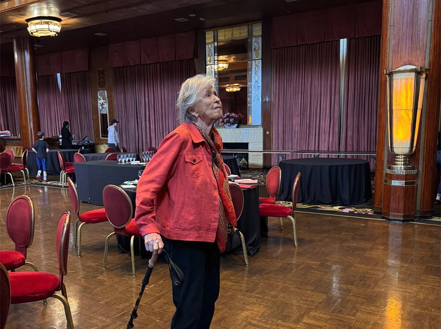 An elderly woman wearing an orange jacket and holding a cane stands in an elegant room with polished wood floors, red chairs, and large windows with pink curtains. Other people are seen in the background.