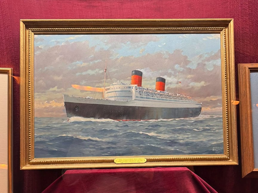 A framed painting of an ocean liner with two red and black smokestacks sailing on choppy waters under a cloudy sky, displayed against a red velvet background.