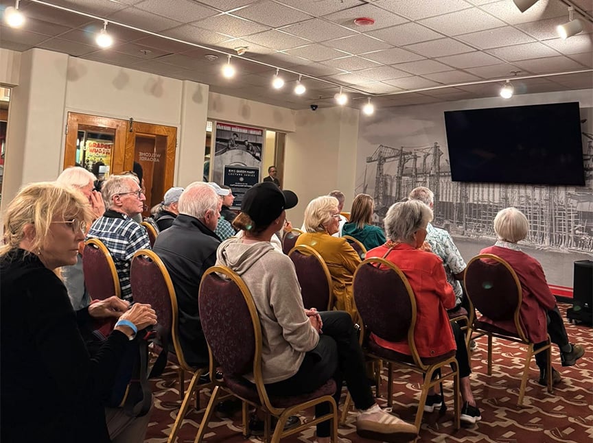 A group of people, mostly older adults, sit on chairs in a room, facing a large blank TV screen on the wall. The room has patterned carpet, overhead lights, and posters on the walls.