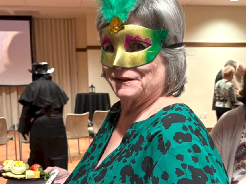 An older woman wearing a green and gold masquerade mask with a feather, dressed in a green patterned top, stands indoors holding a plate of snacks at a social event. Other people are visible in the background.