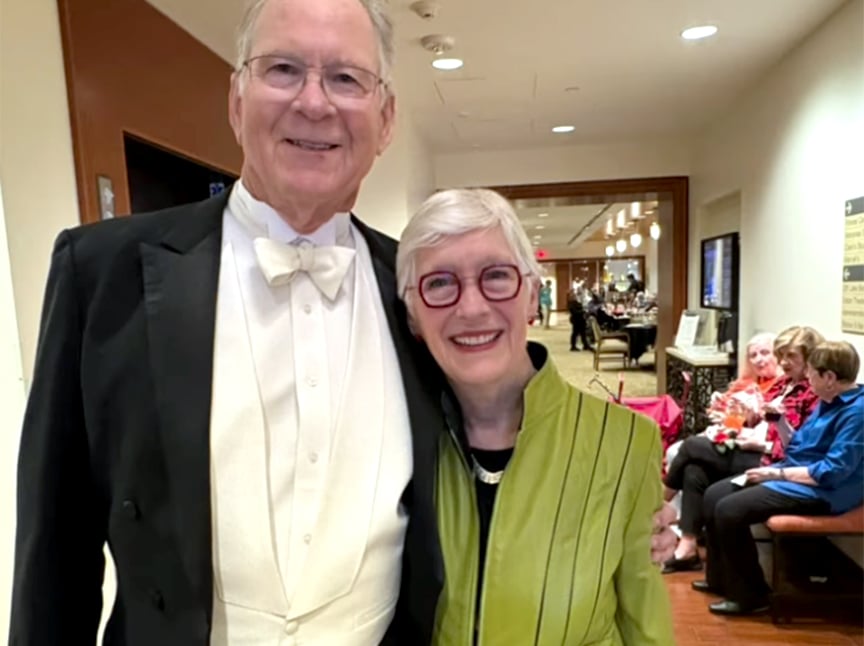 An older man in a black tuxedo with a white bow tie stands smiling beside an older woman in glasses and a light green jacket. They are indoors, with other people seated and talking in the background.