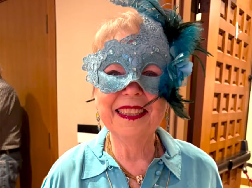 An older woman smiles while wearing a blue lace masquerade mask decorated with feathers and a flower, matching her blue shirt. She has red lipstick and is standing indoors near a wooden door.
