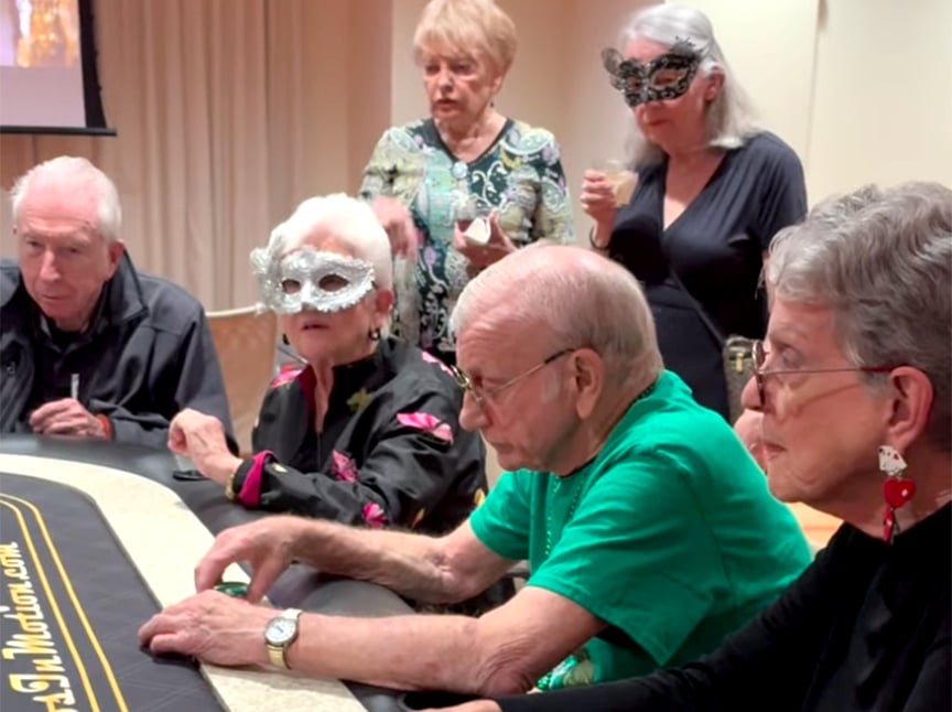A group of elderly people sit and stand around a poker table, some wearing decorative masks. The atmosphere appears festive and social, with attention focused on the game.