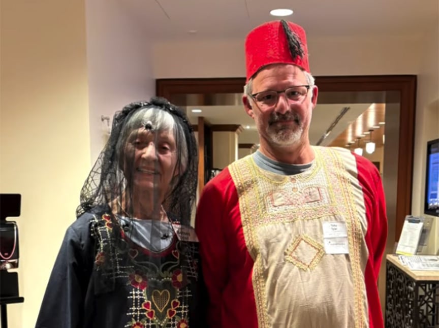 Two people standing indoors wear traditional Middle Eastern attire. The woman has a black embroidered dress and a black veil, while the man wears a red robe with gold embroidery and a red fez hat. Both are smiling for the camera.