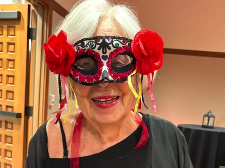 An older woman with white hair smiles while wearing a decorative mask adorned with red roses and ribbons at a festive indoor event.