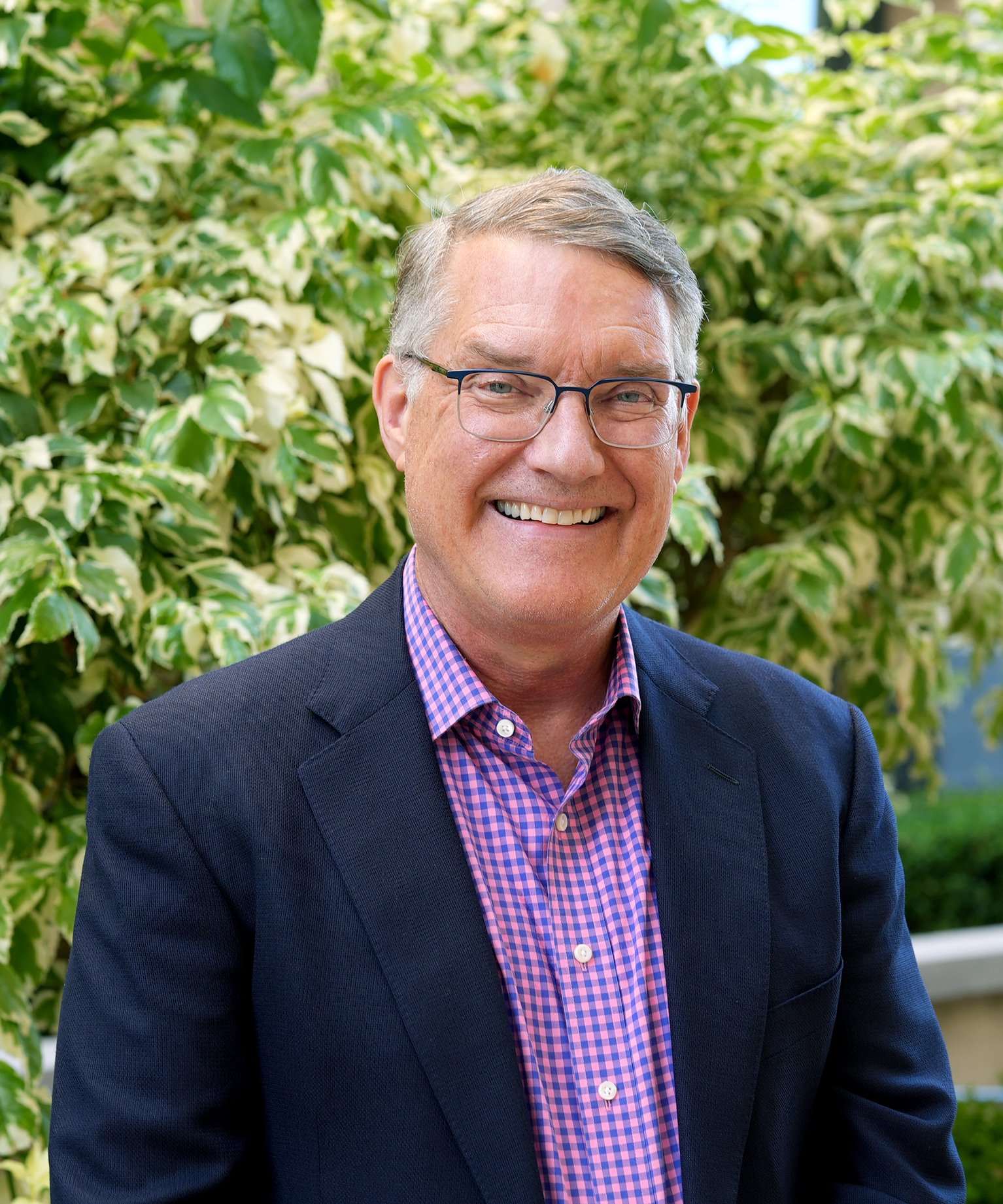 A smiling middle-aged man with gray hair and glasses, wearing a dark blazer over a pink and blue checkered shirt, stands outdoors in front of a variegated leafy plant.