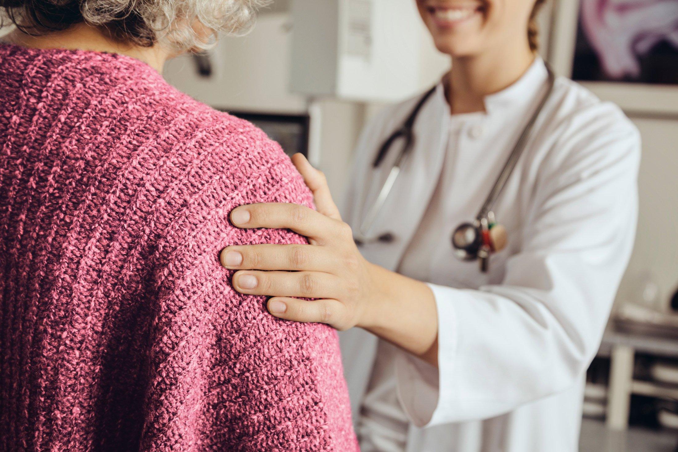 A doctor with a stethoscope smiles and gently rests a reassuring hand on an older patients shoulder, who is wearing a pink knit sweater. The scene conveys comfort and support in a medical setting.