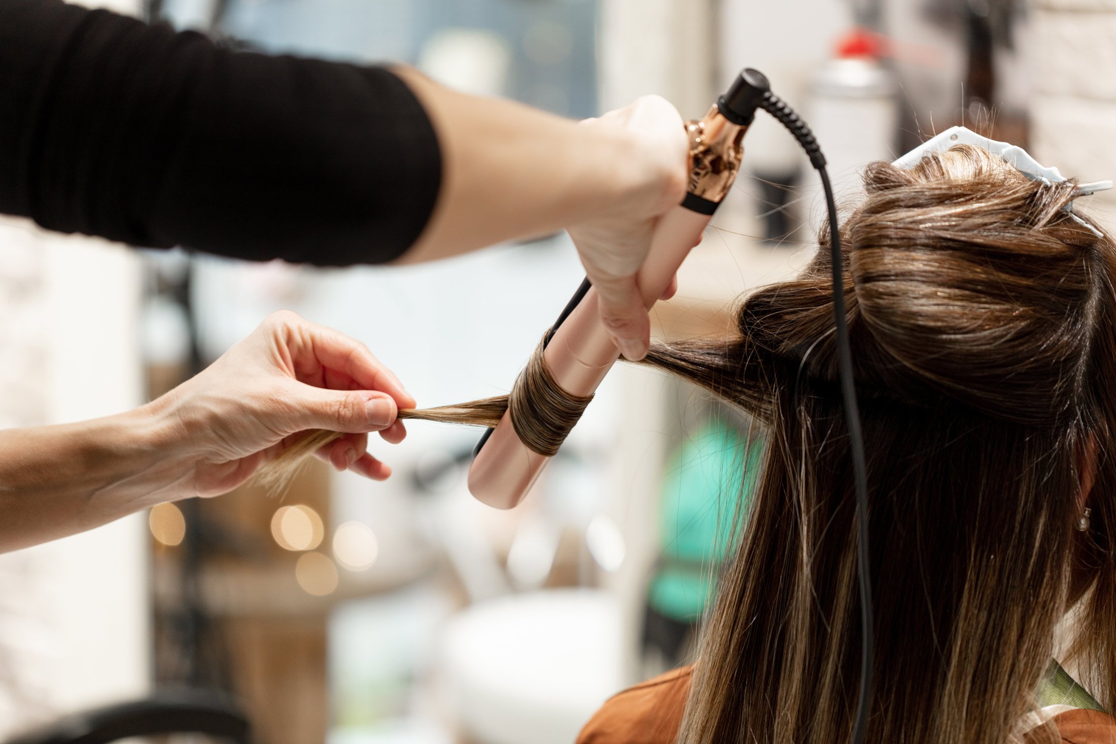 A hairstylist uses a curling iron to curl a section of a womans long, brown hair in a salon. The stylists hands and the curling iron are in focus, with the background softly blurred.