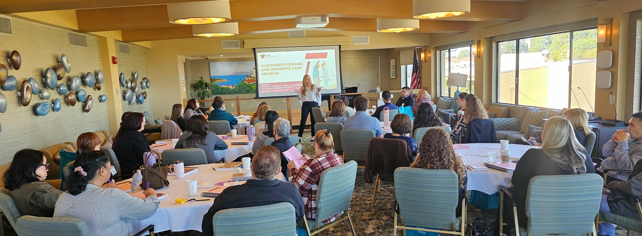 A woman stands and presents to a room full of seated people at round tables. A presentation slide is projected behind her, and attendees are listening and taking notes in a bright, windowed conference room.