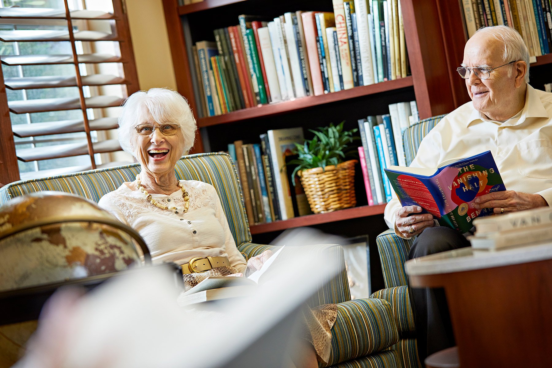 Two elderly people sit and smile in a cozy room with bookshelves. The woman is laughing, and the man is holding a book. Both are seated on striped chairs, and sunlight streams in through a window with wooden blinds.