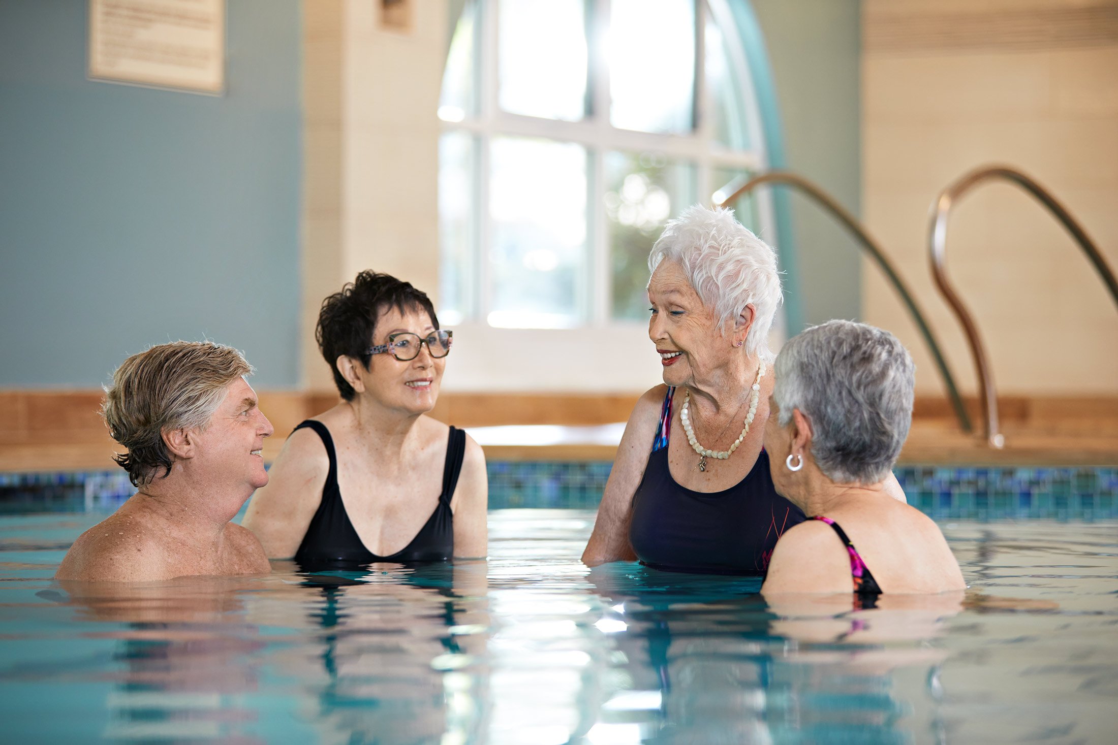 Four older adults stand and talk together in an indoor swimming pool. They are smiling and appear to be enjoying each others company. Sunlight streams through a large window in the background.