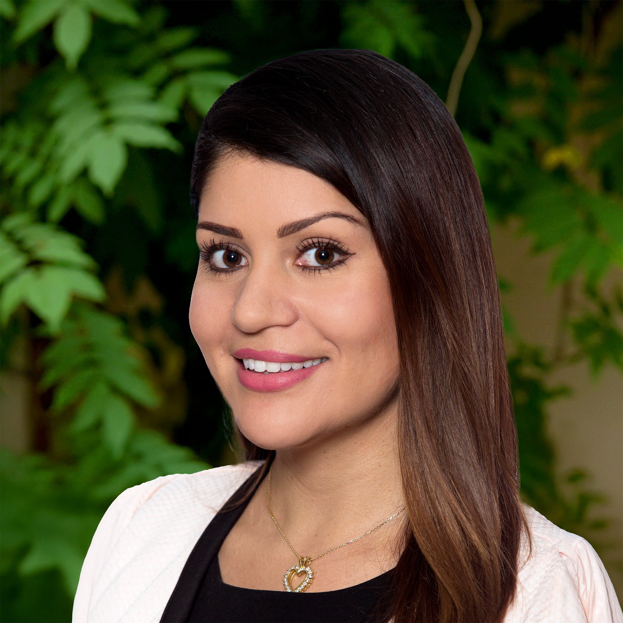 A woman with long, straight dark hair wearing a light-colored jacket and a necklace smiles at the camera. Green leafy plants fill the background.