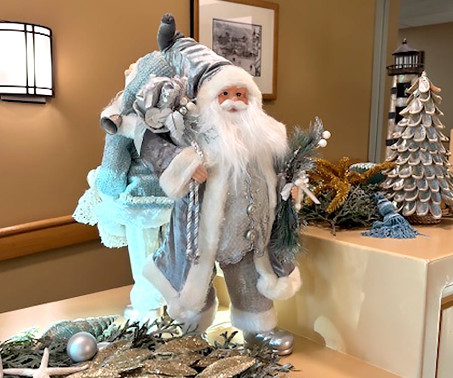 A festive Santa Claus figurine in a silver and white outfit holds greenery and has a sack of gifts on his back. He stands on a beige counter decorated with silver ornaments, stars, and a metallic Christmas tree.