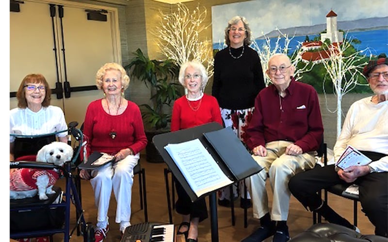 Six smiling elderly people, one with a small white dog, sit indoors with music stands, sheet music, and a keyboard. A painted mural and decorative white branches are in the background.