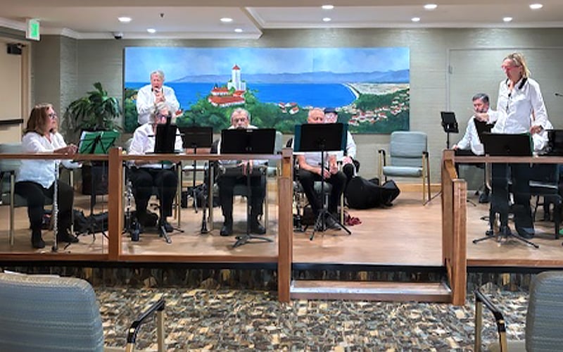 A small band of musicians with instruments sits and stands on a stage indoors, performing in front of an audience. A mural of a coastal landscape with a lighthouse is painted on the wall behind them.