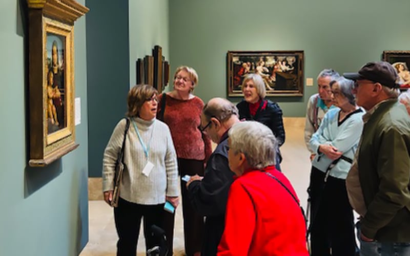 A group of people gather in an art museum, attentively observing and discussing a framed painting on the wall. Several other paintings are displayed in the background.