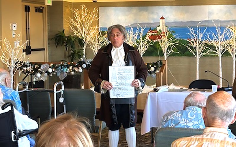 A man in historical costume stands in front of an audience, holding a document. Behind him is a decorated table and a mural of a lighthouse by the sea, with white tree lights and festive garland. Several elderly people are seated and watching.