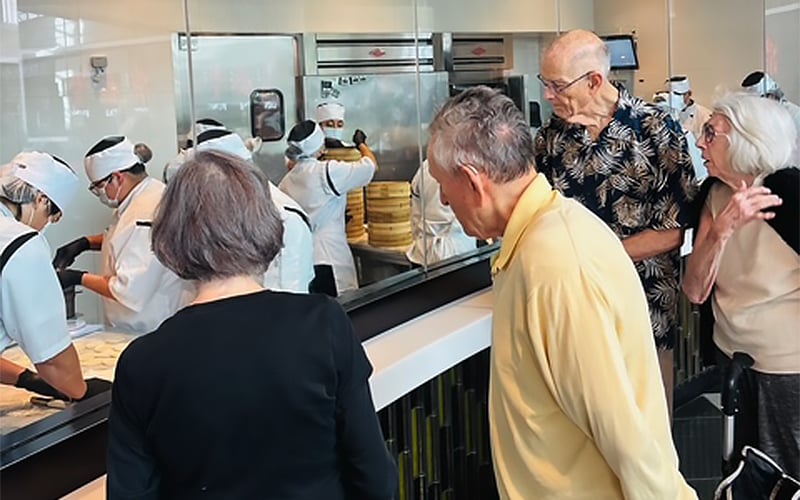 Several elderly people watch through a glass partition as chefs in white uniforms and hats prepare food in a commercial kitchen. The chefs are seen working with steamers and other equipment.