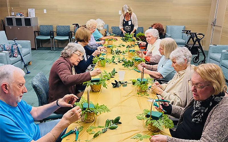 A group of seniors sit around a long table, each working on small plant arrangements. An instructor stands at the head of the table, guiding the activity. The room has comfortable chairs and a calm, communal atmosphere.