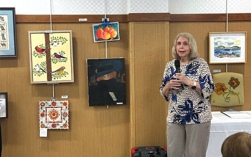 An older woman stands and speaks beside a display of various paintings at an art exhibit. She wears a patterned blouse and holds a microphone. Several colorful artworks hang on the wooden-paneled wall behind her.