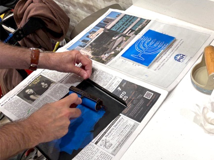 A person uses a roller to spread blue ink onto a surface over newspaper, preparing for printmaking. Nearby, a blue inked design and a photograph of buildings are visible on white paper.