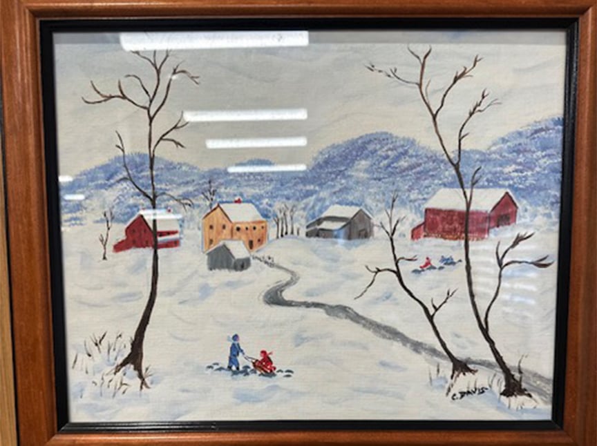A framed painting of a snowy village scene with distant hills, houses, barns, bare trees, and people playing in the snow, including a person pulling a child on a sled along a winding road.