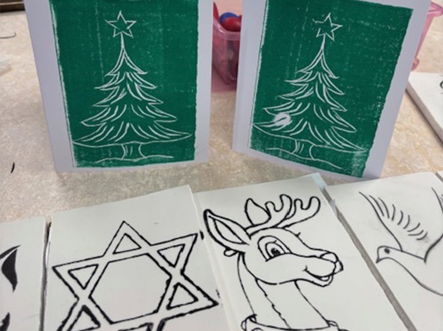 Two handmade holiday cards with Christmas tree prints stand upright. In front of them are black-and-white line drawings of a Star of David, a reindeer, and a dove on white tiles. Art supplies are seen in the background.