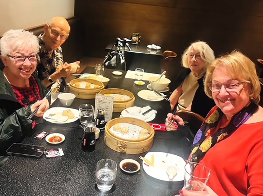 Four smiling older adults sit around a restaurant table enjoying dim sum, with bamboo steamers, soy sauce, and drinks in front of them. They appear happy and are engaged in conversation.