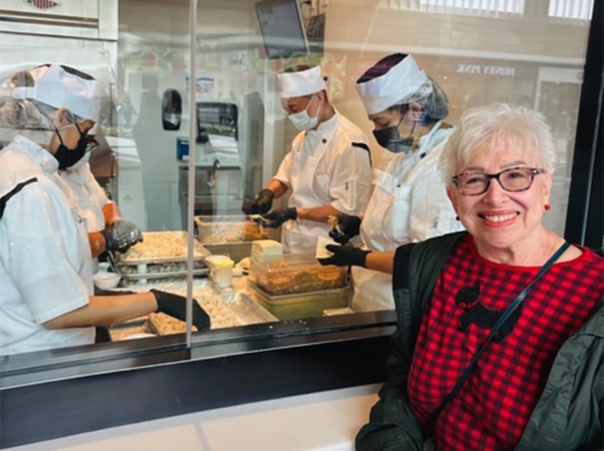 An older woman with short white hair and glasses smiles at the camera. Behind her, several chefs in white uniforms and masks prepare food behind a glass window in a restaurant kitchen.
