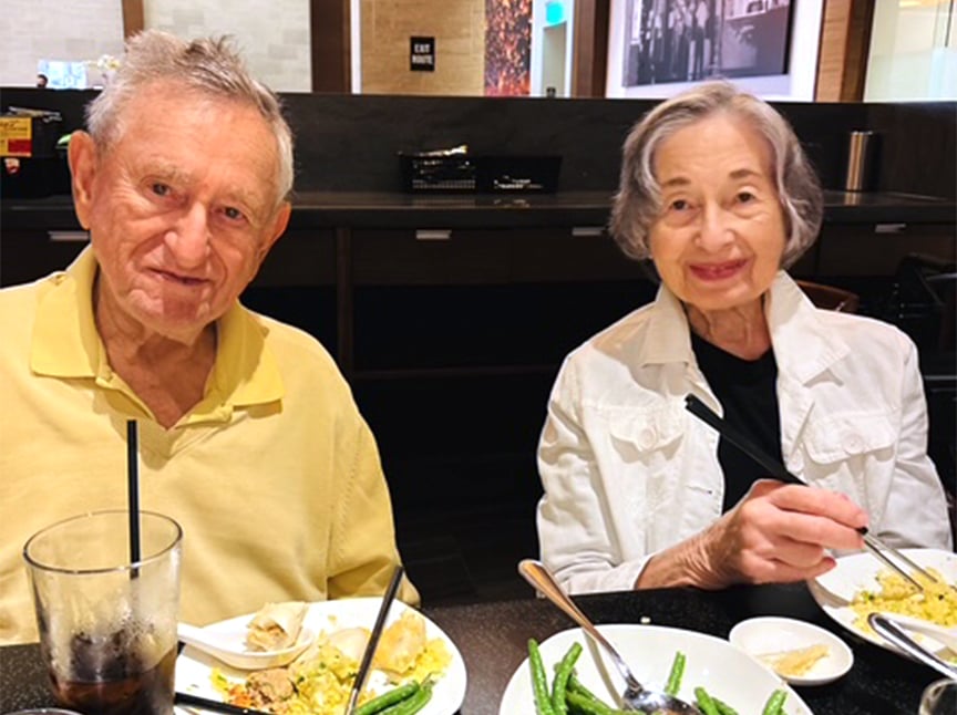 An older man and woman are sitting at a restaurant table, smiling at the camera. They have plates of food and drinks in front of them. The man is wearing a yellow shirt and the woman a white jacket.