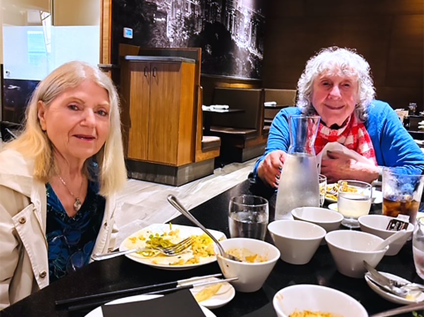 Two older women sit at a restaurant table with several bowls, plates of food, glasses of water, and a pitcher. They are smiling and appear to be enjoying a meal together in a warmly lit dining area.