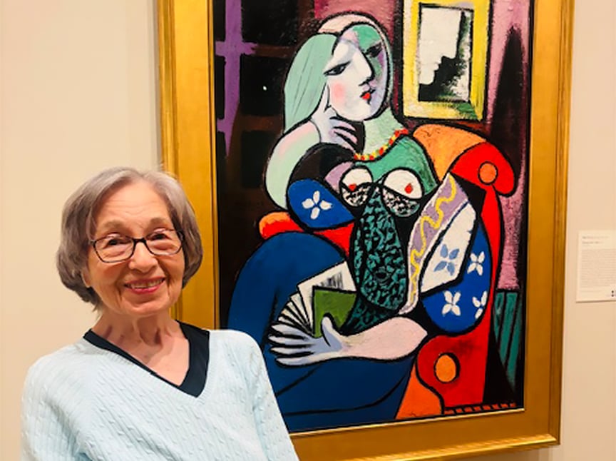 An older woman with gray hair and glasses smiles while standing in front of a colorful, abstract painting of a seated woman in a gold frame at an art gallery.