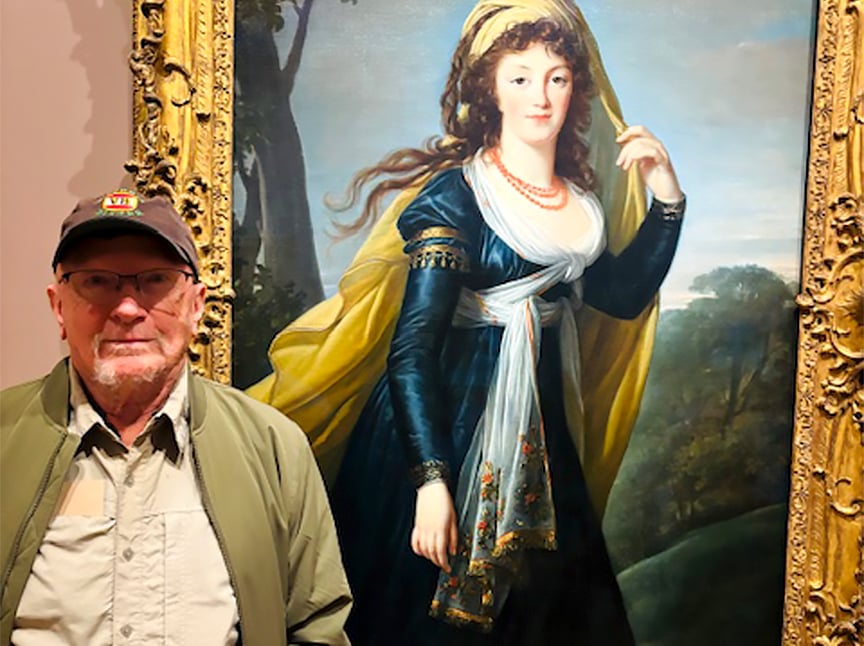 An older man wearing glasses and a cap stands in front of a large, ornate framed painting of a woman in historical dress with a shawl, blue gown, and long hair, posing outdoors.