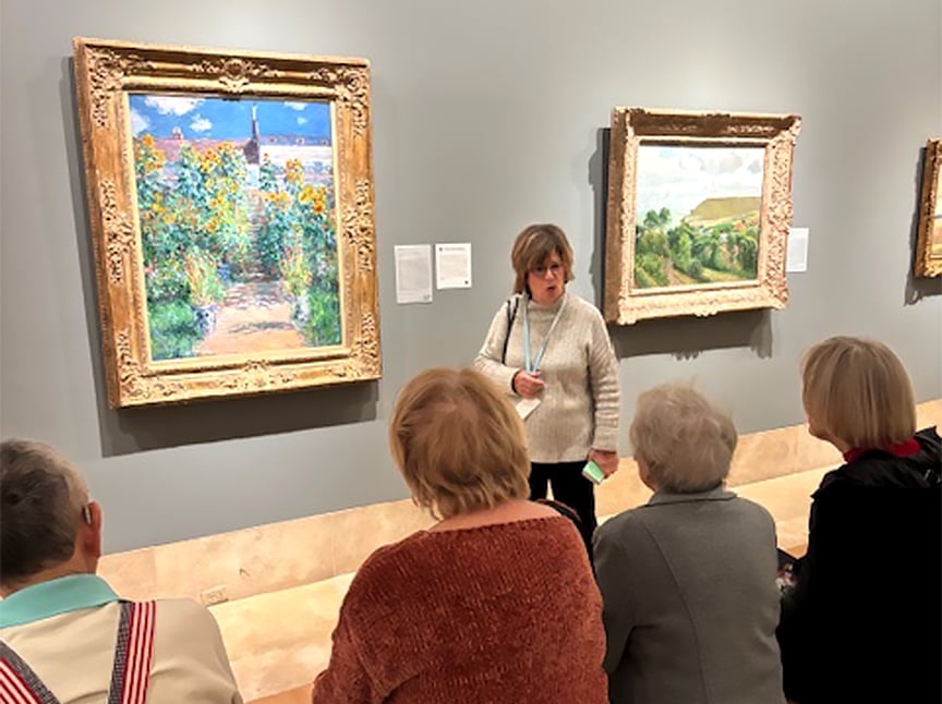 A woman stands in front of a framed painting in an art museum, speaking to a seated group of four older adults who are viewing the artwork and listening attentively.