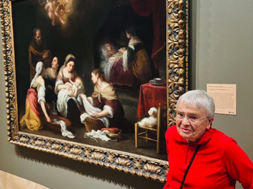 An older woman in a red sweater stands smiling beside a large, ornate-framed painting depicting several people gathered around an infant in a classical setting, displayed in a museum.